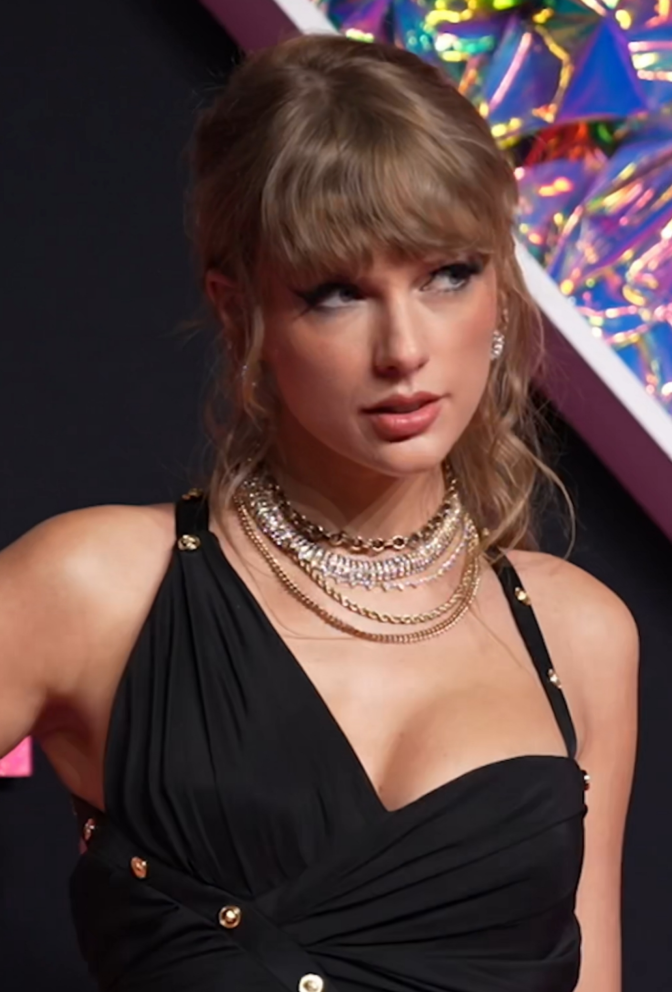 Taylor Swift in einem asymmetrischen schwarzen Glitzerkleid und mit zurückgebundenen Haaren. Sie trägt viel Schmuck und schaut leicht fan der Kamera vorbei aus dem Fenster.