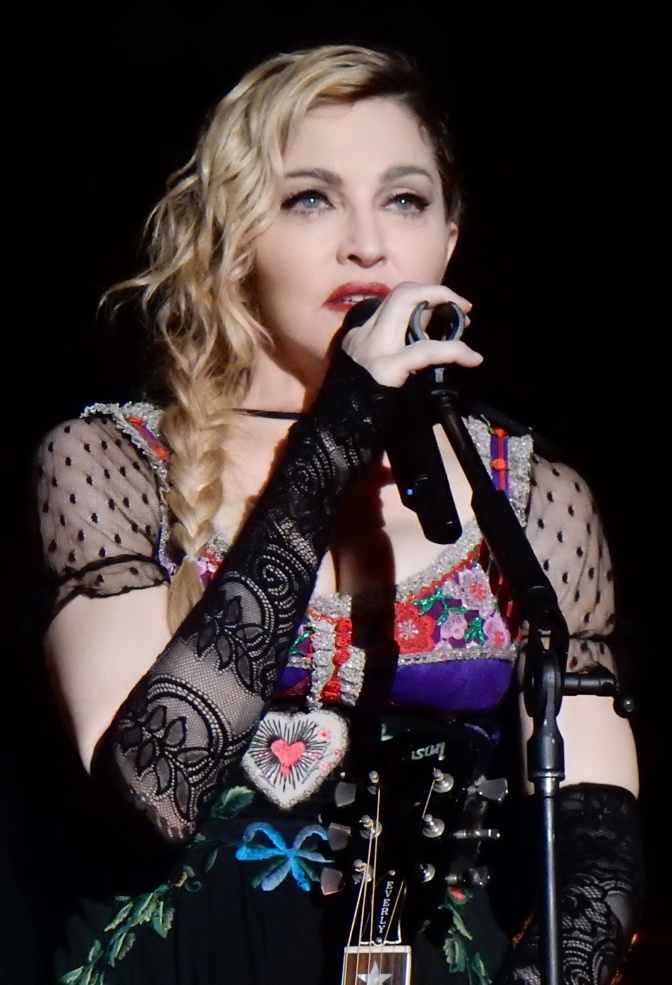 Madonna mit in Wellen gelegten blonden Haaren in einem transparenten schwarzen Bühnenoutfit. Sie singt in ein Mikrofon.