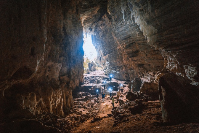 Mehrere Menschen mit Stirnlampen im Eingangsbereich einer großen Höhle. Durch den Höhleneingang fällt Licht ins Innere der Höhle.