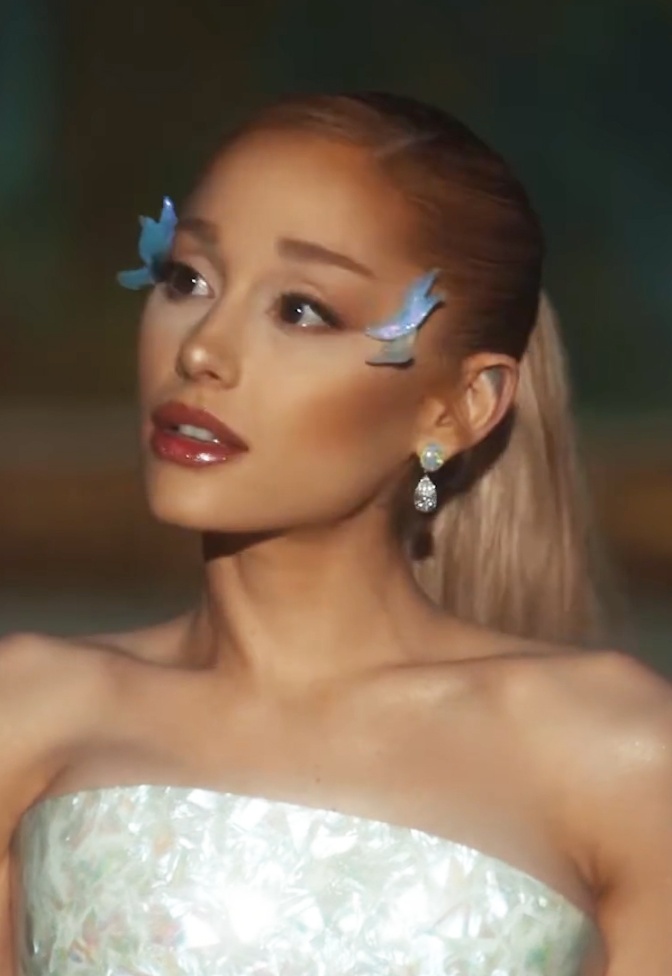 Ariana Grande mit glitzernden Stickern im Gesicht in einem schulterfreien weißen Kleid.