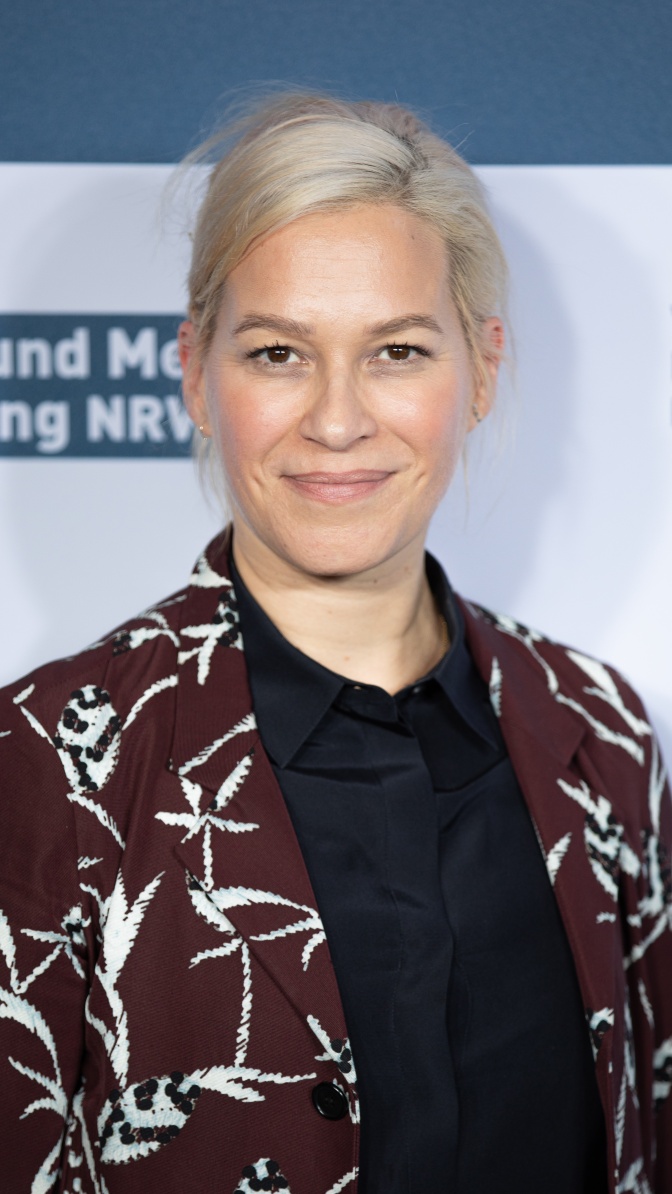 Franka Potente mit blondierten Haaren in einer gemusterten Jacke vor einer Logowand.