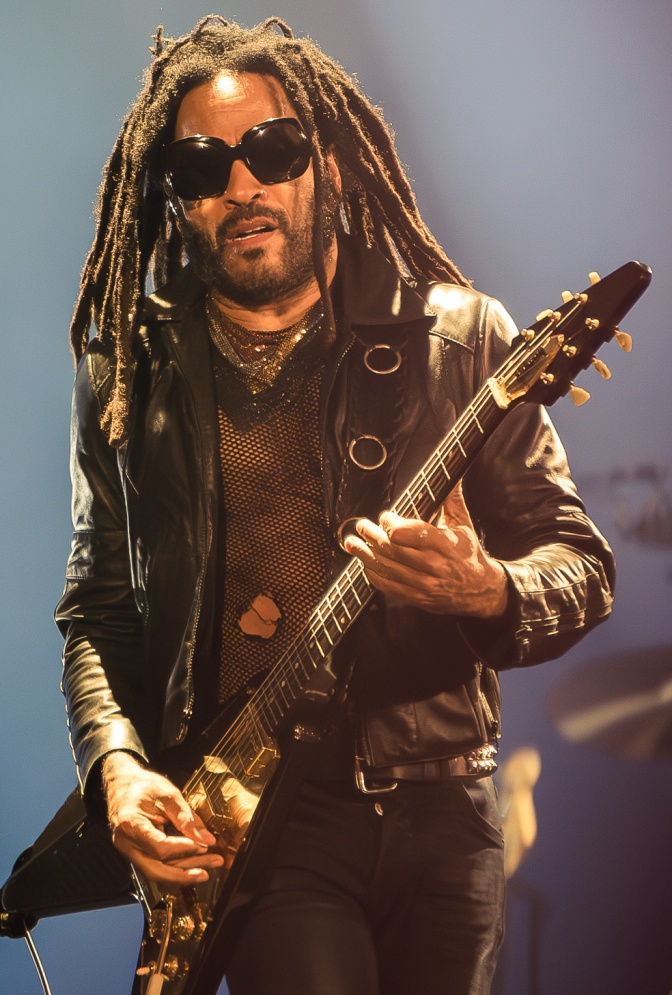 Lenny Kravitz mit langen Dreadlocks und schwarzer Sonnenbrille. Er spielt E-Gitarre und trägt ein Netzshirt.