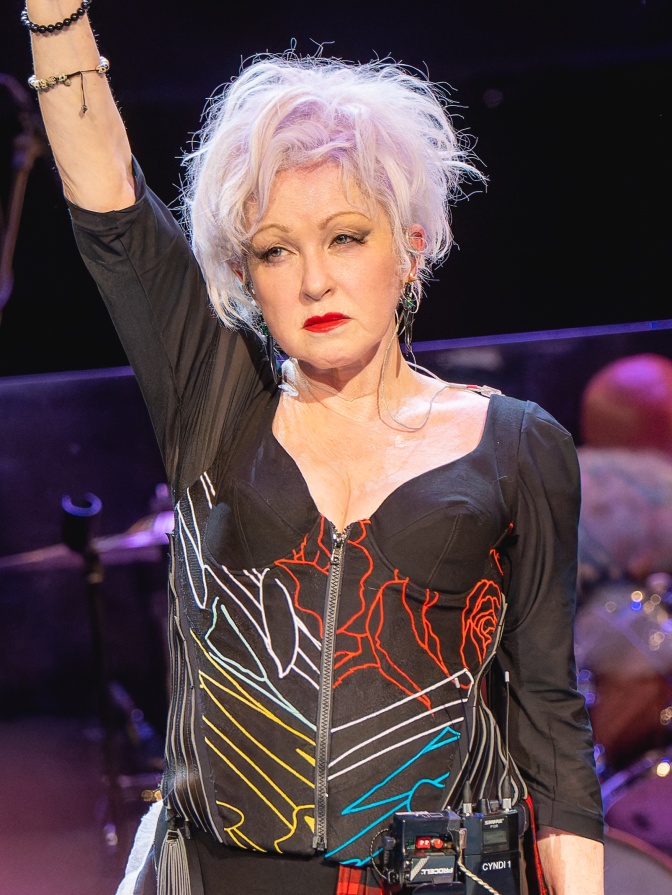 Cyndi Lauper mit blondierten Haaren in einem schwarzen Bühnen-Outfit. Sie streckt einen Arm in die Luft.