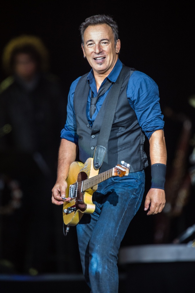 Bruce Springsteen in Jeans und Jeanshemd auf der Bühne. Er hat eine Gitarre an einem Gurt umhängen.