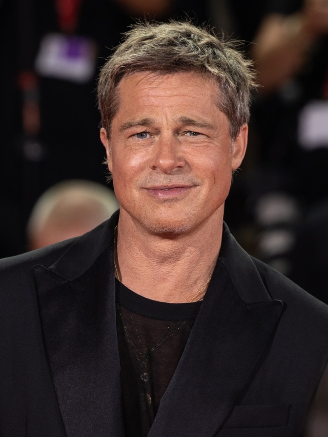 Brad Pitt mit blondierten Haaren in schwarzer Kleidung. Er lächelt.