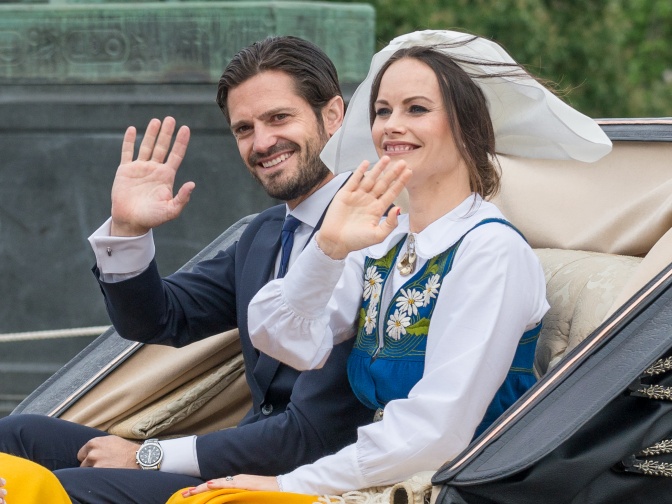 Carl Gustaf und Sofia von Schweden in einer Kutsche. Sie winken. Er trägt Anzug und Krawatte, die schwedische Tracht mit einer weißen Haube.