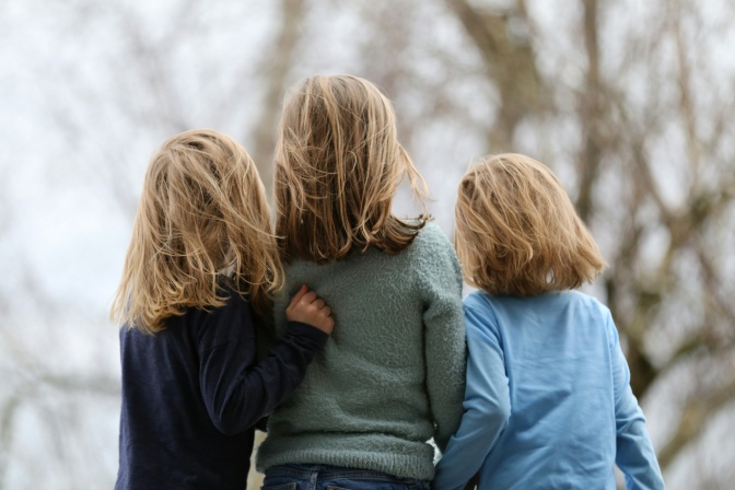 3 Mädchen, von hinten fotografiert. Sie haben schulterlange blonde Haare, die im Wind wehen.