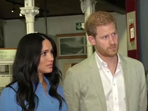 Harry und Meghan in legerer Kleidung. Sie stehen nebeneinander und hören mit ernstem Blick konzentriert zu.