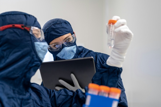 Zwei Personen in Schutzkleidung betrachten ein Reagenzglas, das eine der beiden in der Hand hält.