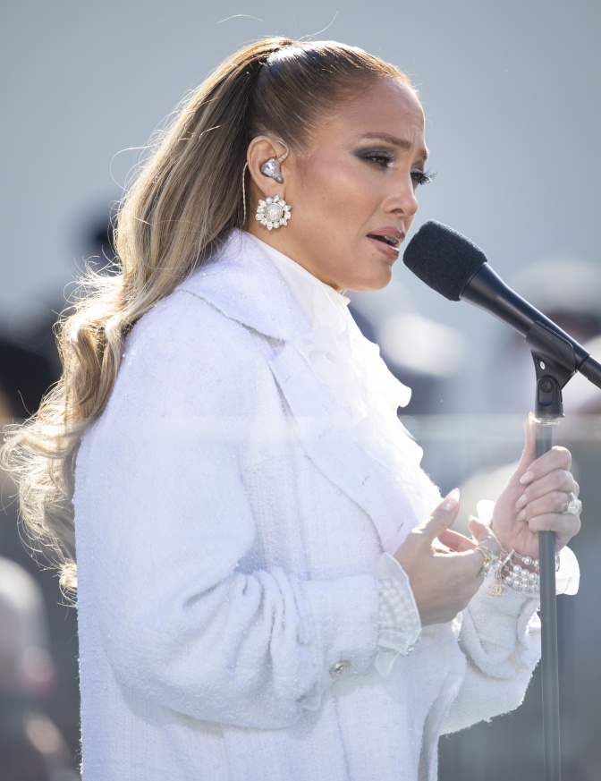 Jennifer Lopez in einem weißen Pelz und mit Pferdeschwanz. Sie spricht in ein Mikrofon.