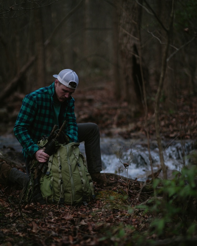 Ein Mann in Karohemd und Schirmmütze sitzt vor einem großen Rucksack im Wald.