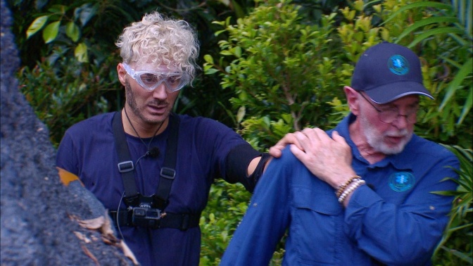 Ein Mann mit blonden Locken mit einer Schwimmbrille folgt einem Mann in blauem Overall. Er hat seine Hand auf seiner Schulter liegen.