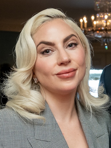 Lady Gaga mit in Wellen gelegten blondierten Haaren in einem grauen Blazer. Sie hat den Kopf schräg gelegt und schaut direkt in