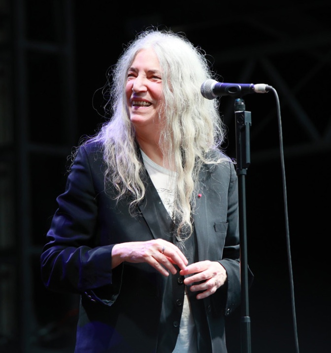 Patti Smith mit langen, weißen Haaren. Sie steht lächelnd in schwarzer Kleidung an einem Standmikro.