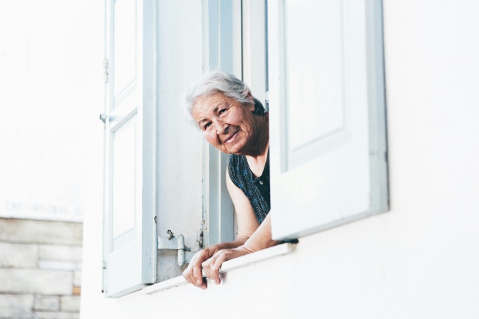 Eine alte Frau mit kurzen weißen Haaren schaut aus einem Fenster heraus.