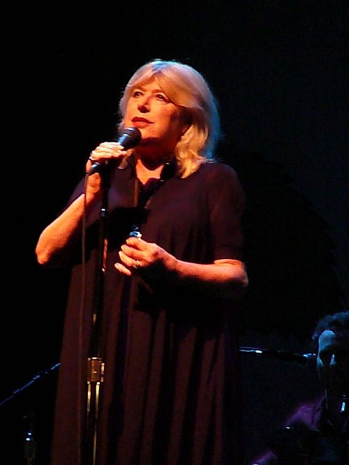 Marianne Faithfull in schwarzer Kleidung auf der Bühne. Sie hält ein Mikro in der Hand und singt.