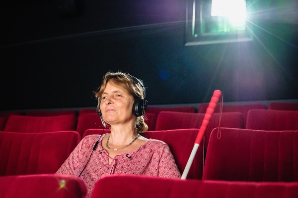 Eine Frau sitzt mit Kopfhörern im Kino. Am Sitz neben ihr lehnt ihr Blindenstock.