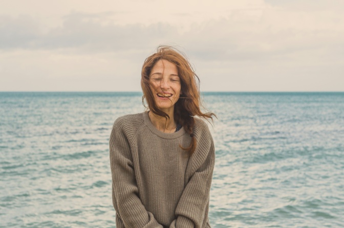 Eine Frau steht am Meer. Der Wind zerzaust ihr schulterlanges Haar. Sie trägt einen dicken, hellbraunen Wollpullover.