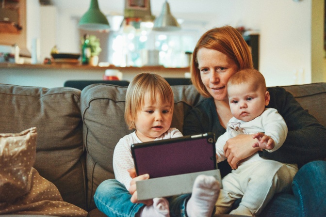 Eine Mutter mit 2 Kindern sitzt auf der Couch und alle 3 schauen auf das Display eines Tablets.