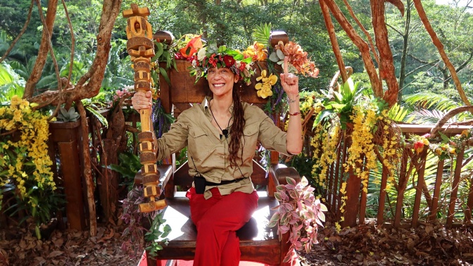 Lilly Becker sitzt mit einem Blumenkranz auf dem Kopf auf einem Thron. Sie hält einen Holzstab in einer Hand und zeigt mit der anderen Hand nach oben.