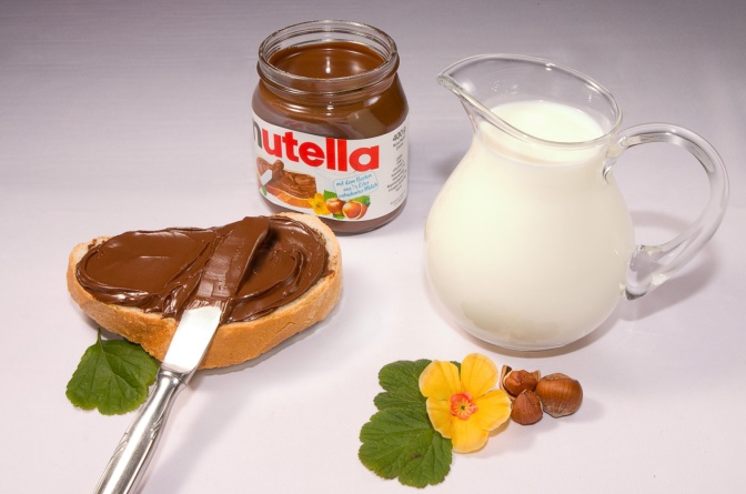 Ein Glas Nutella, daneben ein mit Nutella bestrichenes Brot und ein Krug mit Milch.