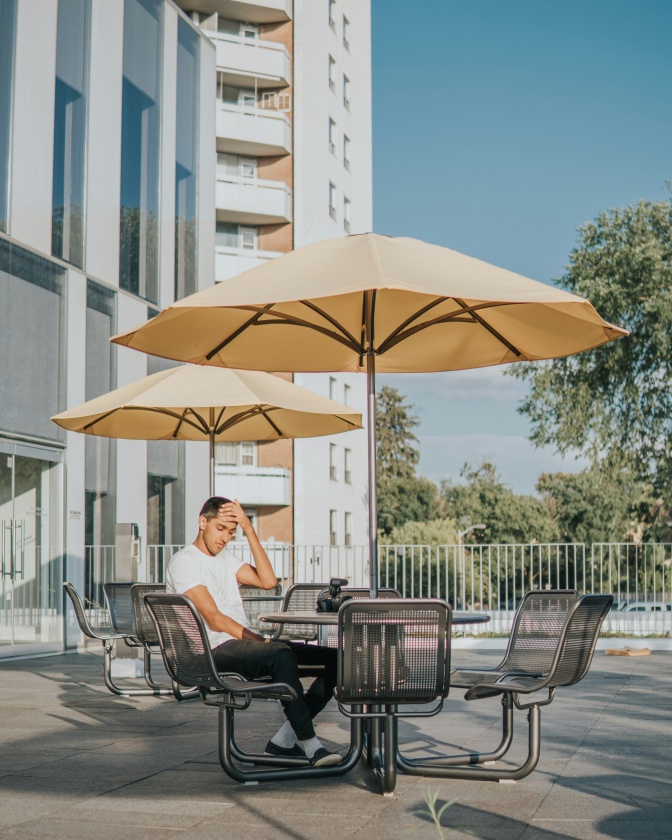 Ein Mann sitzt im Freien an einem Metalltisch unter einem Sonnenschirm. Er verdeckt einen Teil seines Gesichts mit der Hand.