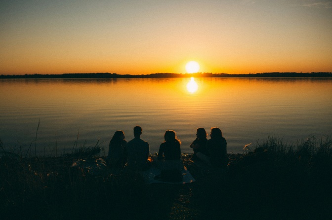 5 Menschen sitzen bei Sonnenuntergang an einem See und unterhalten sich. Man sieht nur ihre Körper von hinten im Gegenlicht.