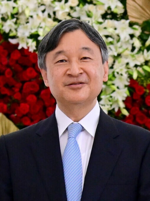 Kaiser Naruhito mit kurzen, grau werdenden Haaren. Er steht vor einer mit Blumen geschmückten Wand.