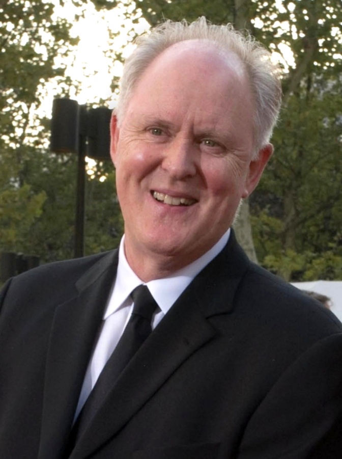 John Lithgow in Anzug und Krawatte. Er hat Stirnglatze und einen weißen Haarkranz. Er steht im Freien und lächelt in die Kamera.