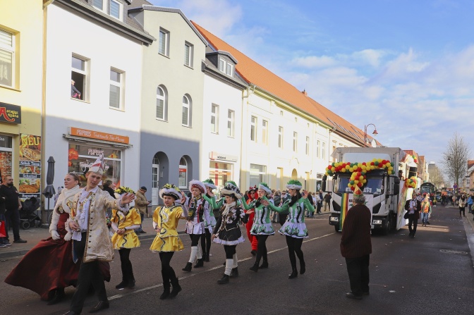 Menschen in Kostümen und Karnevalsuniformen ziehen durch die Straßen und winken den Menschen am Straßenrand zu. Hinter einer Fußgruppe fährt ein mit Luftballons geschmückter LKW.