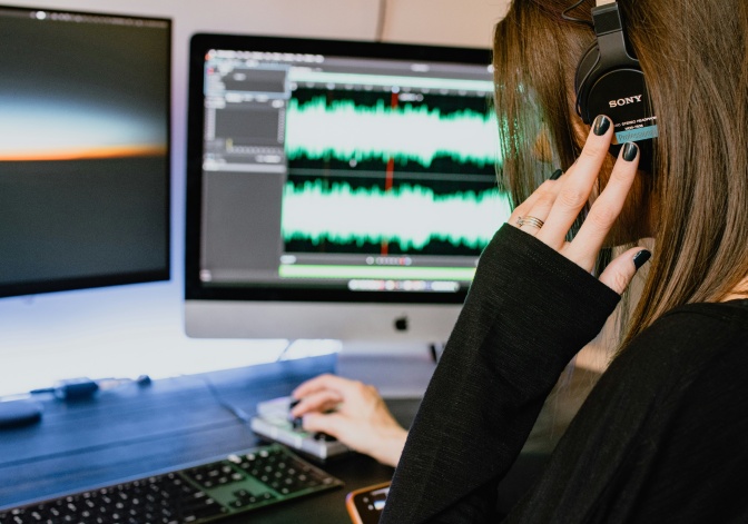 Eine Frau hat Kopfhörer auf und regelt mit der Maus Tonspuren am Computer.