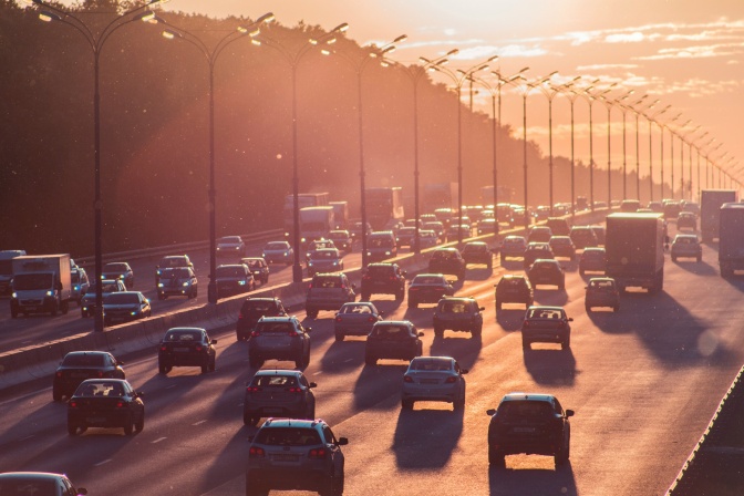 Sich stauende Autos auf zwei vierspurigen Straßen. Der Himmel ist vom Sonnenuntergang orange gefärbt.