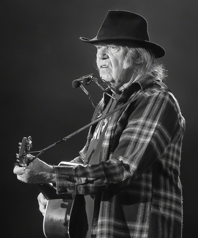 Schwarz-weiß-Foto von Neil Young mit Hut und Karohemd. Er spielt Gitarre, eine Mundharmonika klemmt in einem Gestell vor seinem Mund.