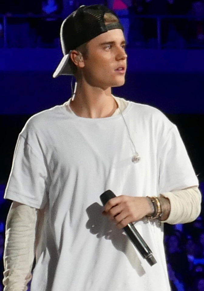 Justin Bieber in einem weißen Shirt und einer nach hinten gedrehten schwarzen Schirmmütze. Er steht auf der Bühne und hält ein Mikro in der Hand.