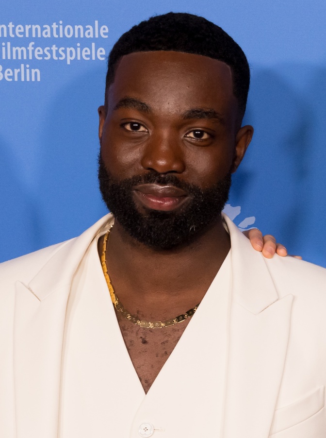 Paapa Essiedu in weißer Kleidung vor einer Logowand der Filmfestspiele in Berlin. Er hat sehr kurz rasierte Haare und trägt Vollbart.