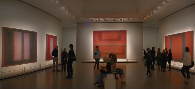 Große Bilder in dunklen Rottönen hängen in einem Museumsraum. Besucher*innen bewegen sich durch den Raum und betrachten die Kunst.
