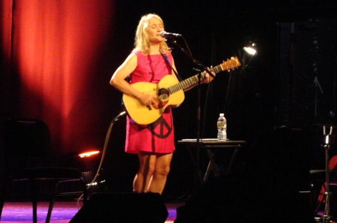 Jill Sobule steht in einem roten Kleid und mit einer akustischen Gitarre auf der Bühne.