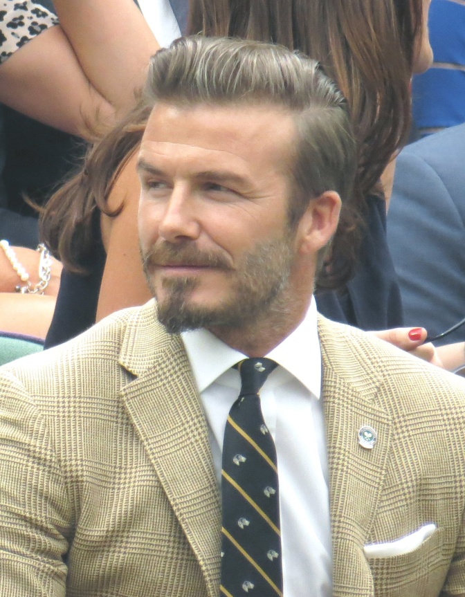 David Beckham mit Vollbart und mit Pomade frisierten Haaren. Er trägt Anzug und eine gestreifte Krawatte und sitzt im Publikum einer Veranstaltung.