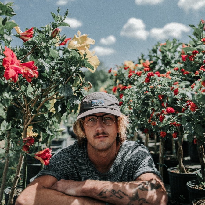 Ein Mann mit Brille, Schnäuzer und blondierten Haaren sitzt in einem Blumenbeet. Er hat Tattoos an den Armen und schaut direkt in die Kamera.