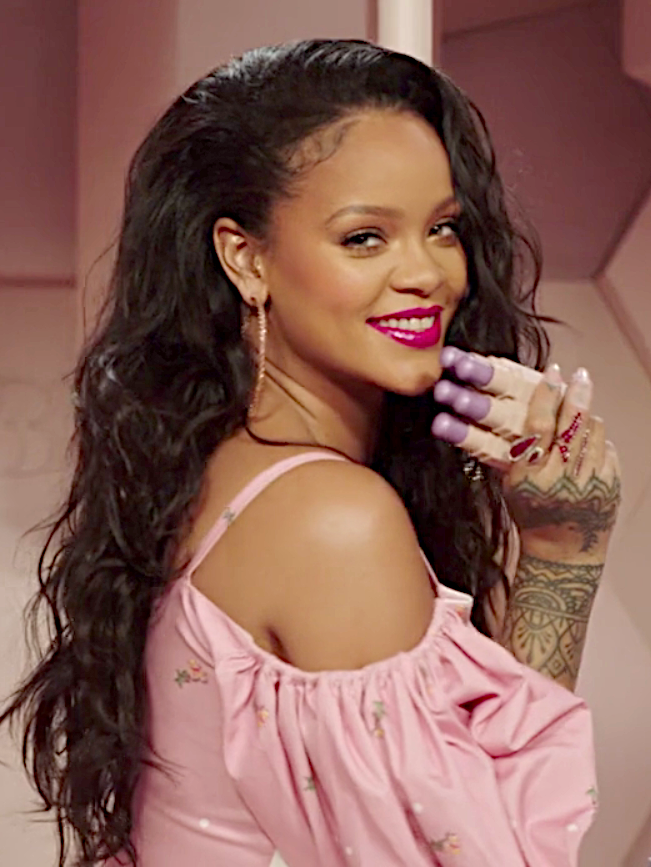 Rihanna in einer rosafarbenen Bluse. Sie hält mehrere fliederfarbene Lippenstifte in der Hand und lächelt über die Schulter in die Kamera.
