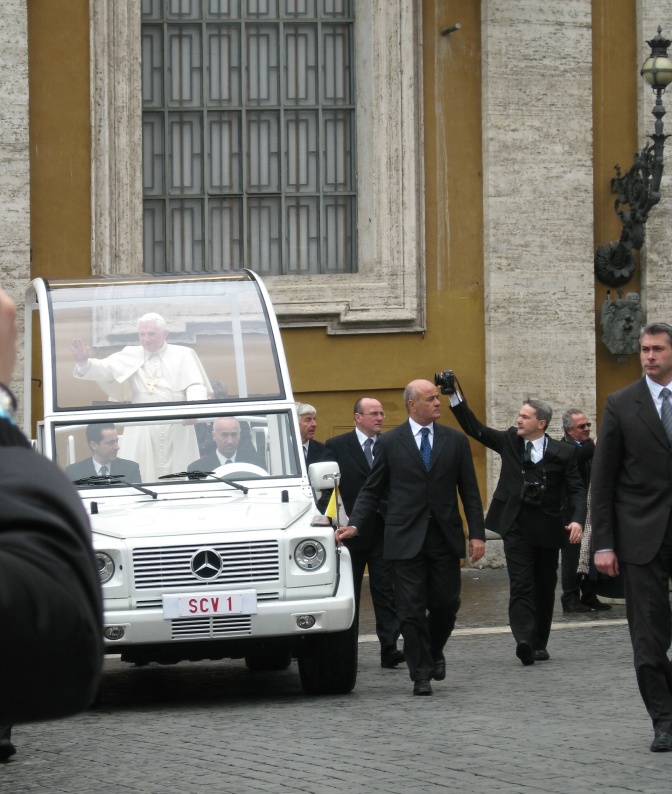 Der Papst wird stehend in einem weißen Auto mit einem durchsichtigen kumpelförmigen Aufbau gefahren. Er winkt Menschen um das Auto herum zu.