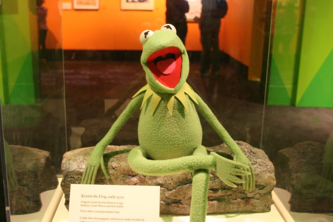 Eine Kermit-Puppe in einer Ausstellungsvotrine. Er sitzt im Schneidersitz und hat den Mund weit aufgerissen.