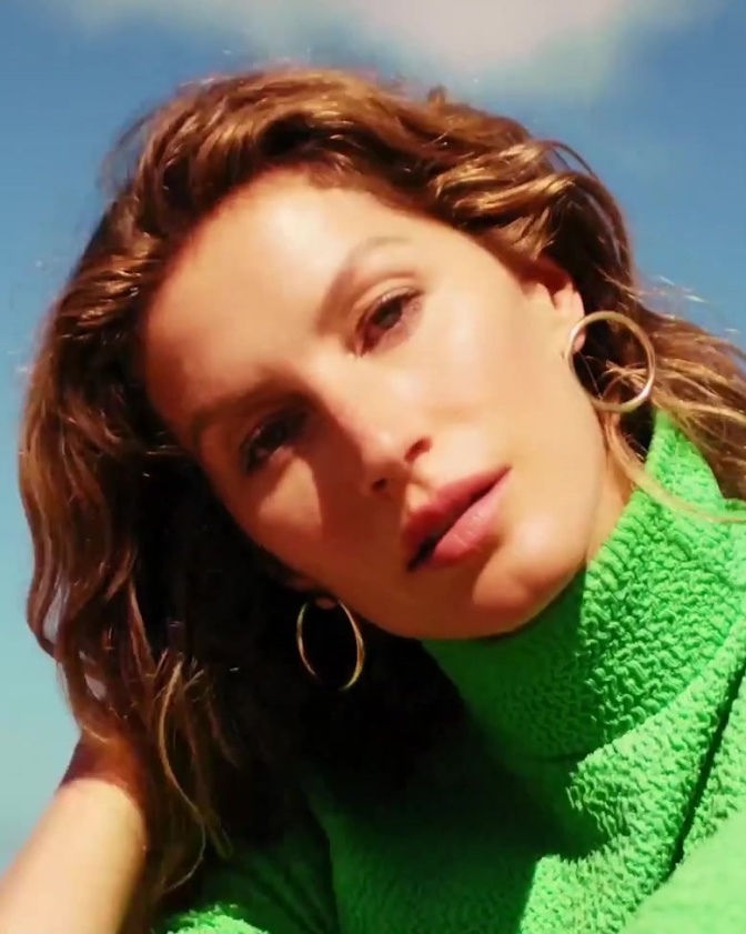 Gisele Bündchen schaut mit schräg gelegtem Kopf direkt in die Kamera. Sie trägt ein grünes Oberteil mit großem Rollkragen.