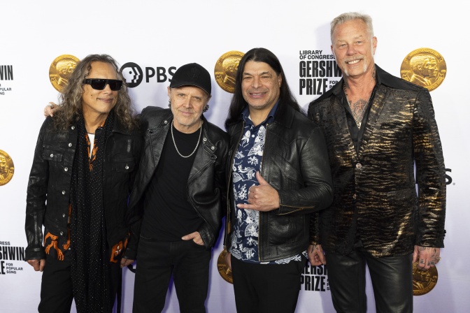 Die 4 Mitglieder der Band Metallica stehen in schwarzer Lederkleidung nebeneinander vor einer Logowand.