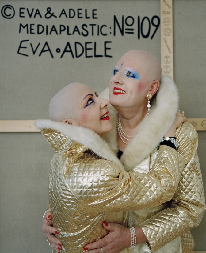 2 Personen mit Glatze und starkem Make up in goldener Kleidung. An der Wand steht: Eva und Adele.