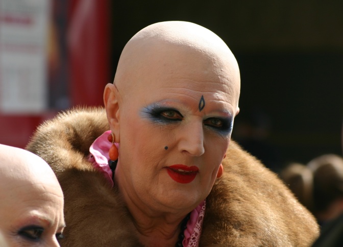 Eine Person mit Glatze und starkem Make up. Die Person trägt Pelz und auffällige Ohrringe.