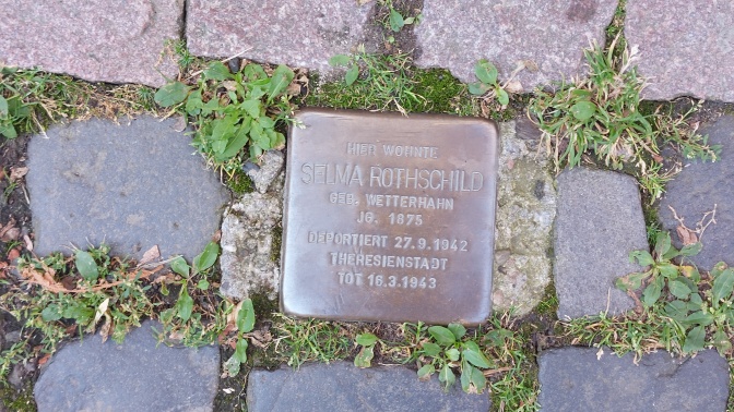 Ein quadratischer kupferfarbener, beschrifteter Stein, der ins Kopfsteinpflaster eingelassen ist. Er ist von Gras und Moos umwachsen.