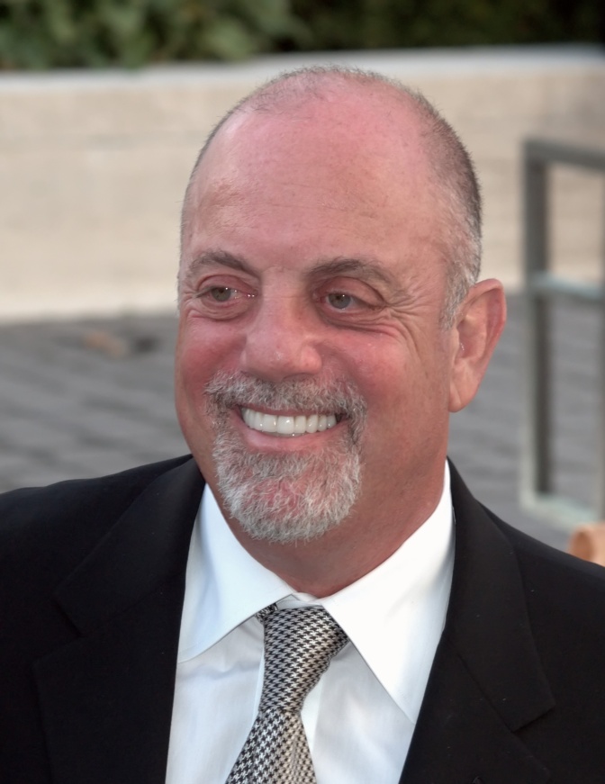 Billy Joel mit Stirnglatze, Vollbart und Krawatte. Er lächelt breit.