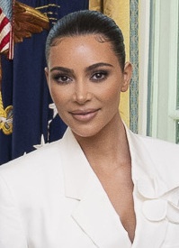 Kim Kardashian in einem weißen Blazer mit streng zurückgebundenen Haaren vor der amerikanischen Flagge.