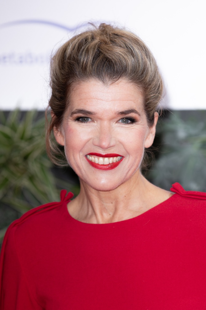 Anke Engelke mit zurückgebundenen welligen Haaren in einem roten Pullover und rotem Lippenstift. Sie lächelt direkt in die Kamera.